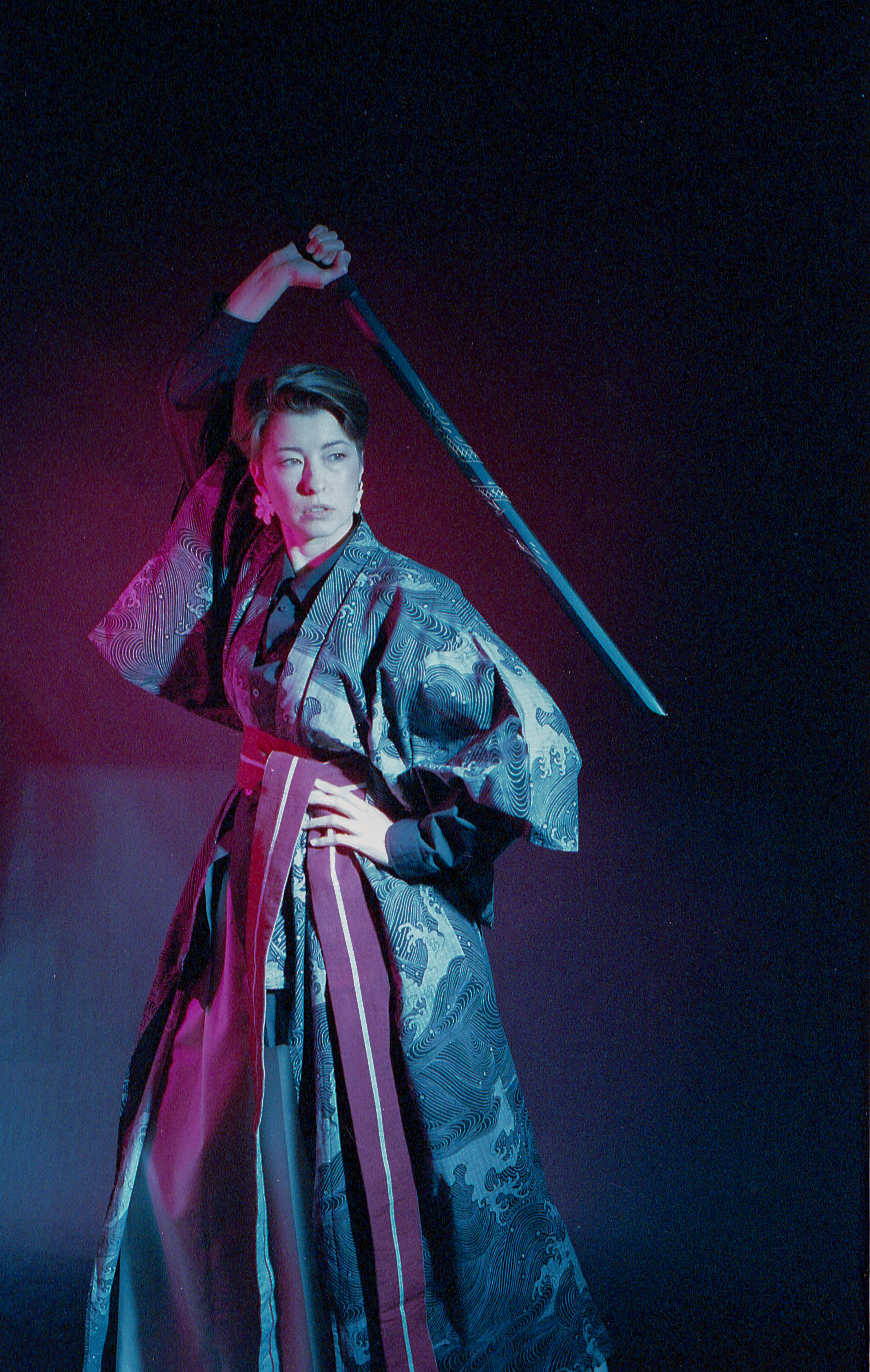 Yulia mid-stance, katana raised, striped hakama in pink light